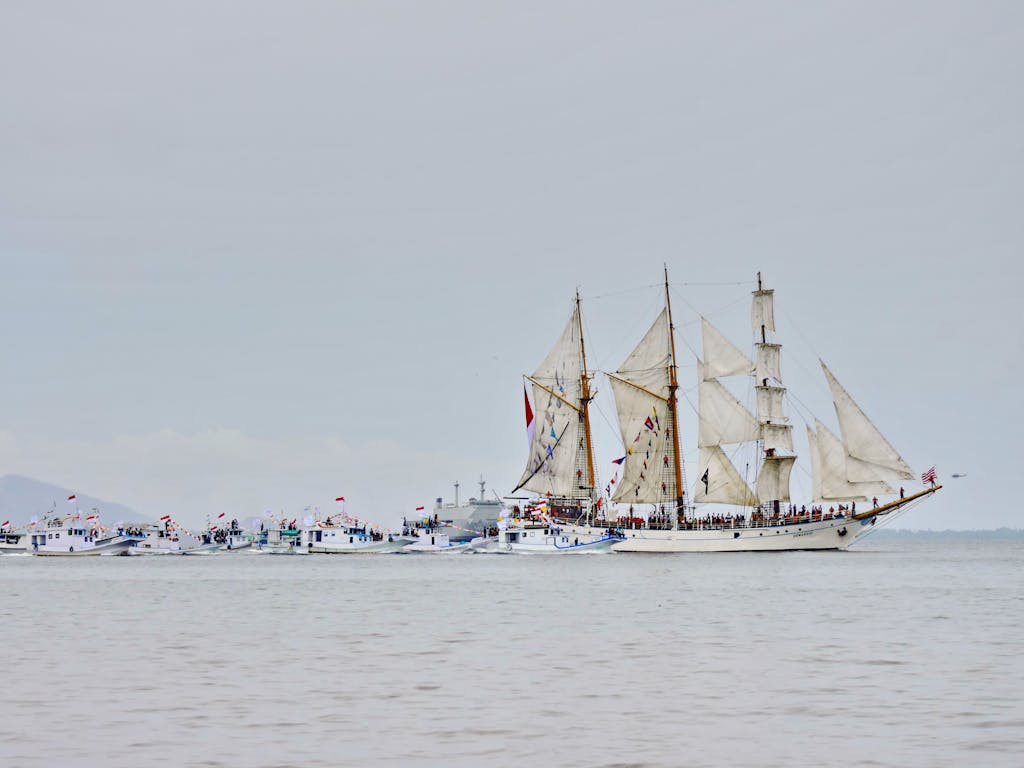 A grand sailing ship with its sails fully unfurled, leading a fleet of boats on a calm sea.