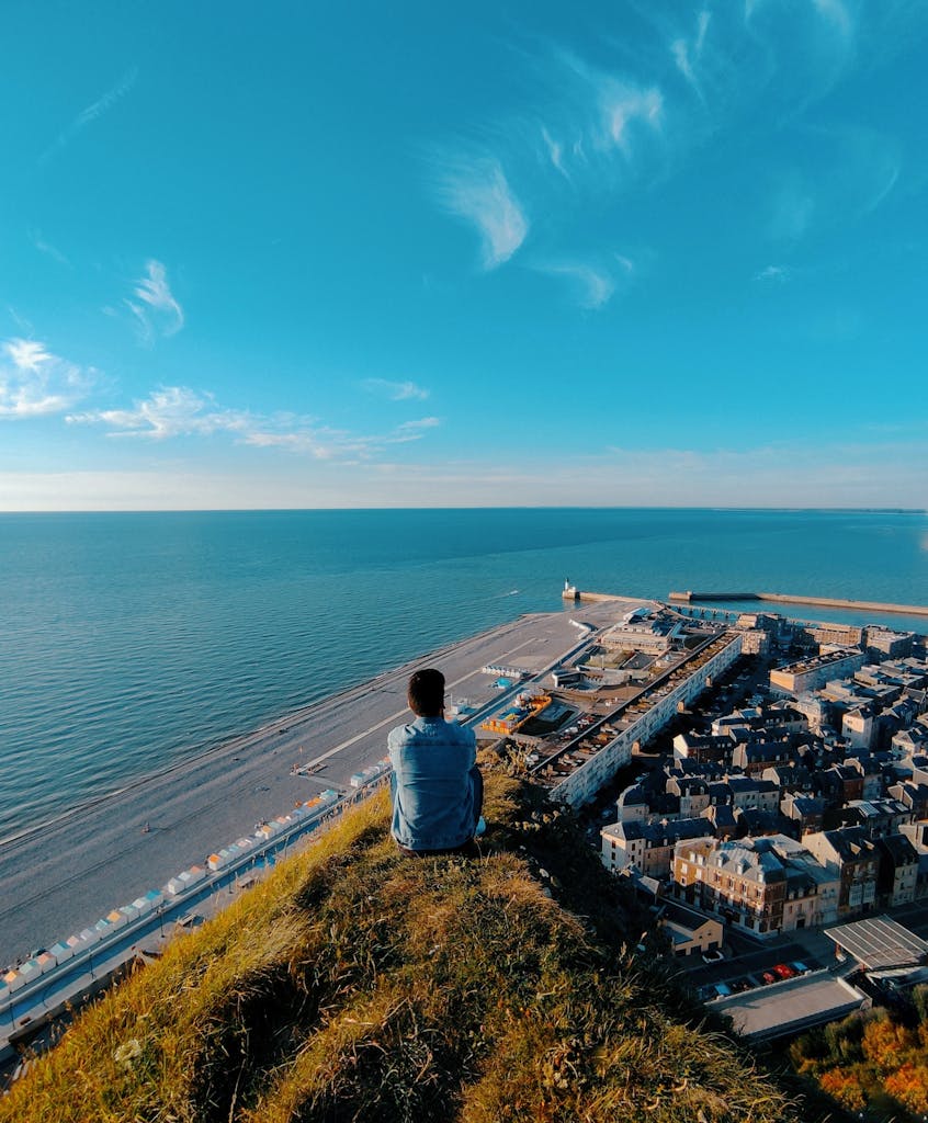 A man sits on a cliff at Le Tréport, Normandy, with a scenic view of the town and sea.