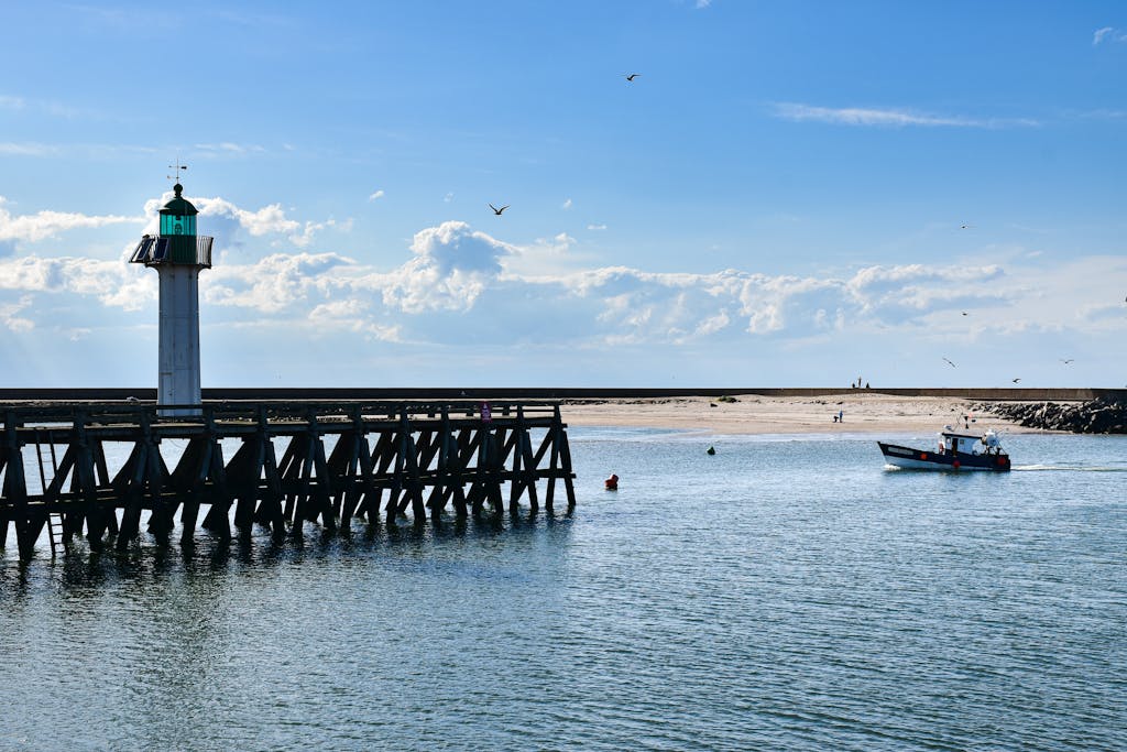 Lighthouse and fishing boat in Honfleur Harbor, Normandy, under a bright blue sky.