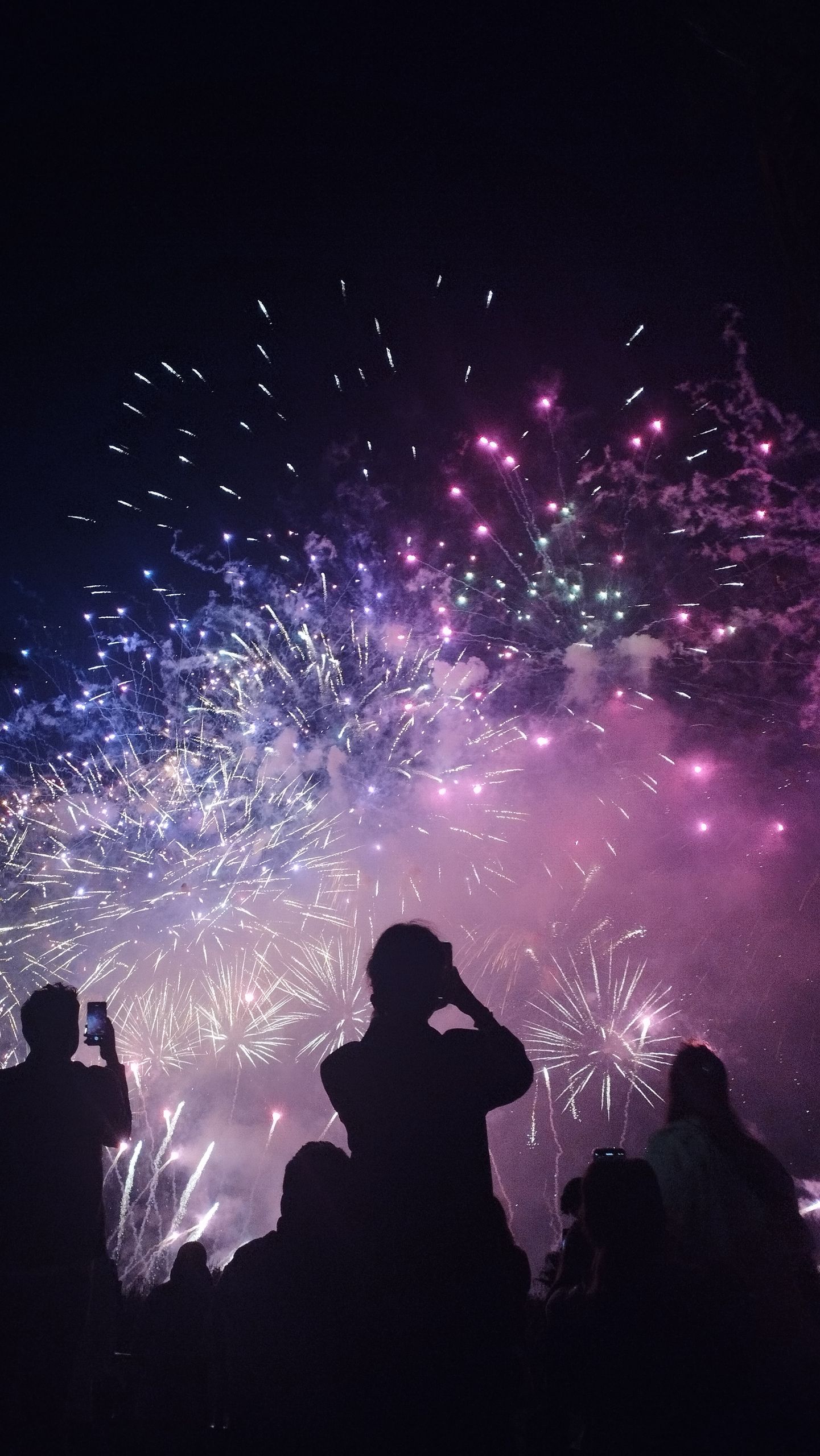 Crowd silhouetted against vibrant fireworks lighting up the night sky, capturing the moment with phones.